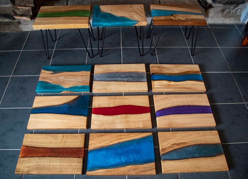 May include: A collection of 10 wooden serving trays with various colored resin river designs. The trays are arranged in three rows, with the top row featuring three trays on black hairpin legs. The resin designs include blue, green, gray, red, purple, and brown.