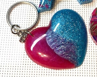 May include: A heart-shaped keychain with a silver keyring. The heart is split diagonally, with a vibrant pink on the bottom and a sparkling blue on top. The blue section has white glitter. Other resin pieces are visible in the background.