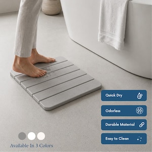 May include: A gray, rectangular bath mat with a slatted design. The mat is shown with bare feet standing on it near a white bathtub. The mat is available in three colors and is described as quick-drying, odorless, durable, and easy to clean.