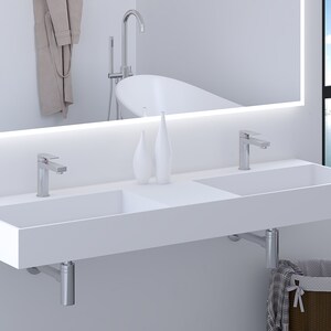 May include: A modern white double sink with two chrome faucets. The sink is mounted on the wall and has a minimalist design. A large mirror with a light strip is above the sink. A bathtub is reflected in the mirror.