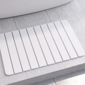 May include: A white rectangular bath mat with a slatted design. The mat is made of a solid white material and is positioned on a light gray tiled surface. The mat is placed near a white bathtub.