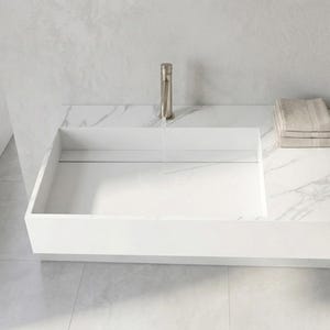 May include: A modern white rectangular sink with a brushed nickel faucet. The sink is integrated into a countertop with a marble pattern. A stack of beige towels sits on the countertop. The bathroom fixtures are set against a light gray wall.