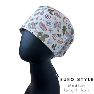 May include: A Euro-style scrub cap with a cream background and a repeating pattern of pie slices, flowers, leaves, and crescent moons. The cap is displayed on a black mannequin head. The text "EURO-STYLE" and "Medium length hair" is visible.