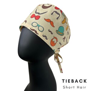 May include: A beige tieback surgical scrub cap with a repeating pattern of mustaches, bow ties, eyeglasses, and hats in various colors. The cap is tied at the back and is designed for short hair. The text "TIEBACK Short Hair" is visible.