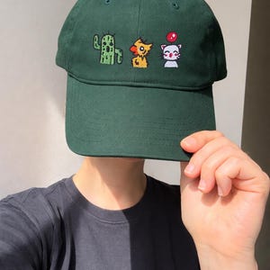 Puede incluir: Gorra de béisbol verde oscuro con bordado de pixel art de un cactus, una criatura parecida a un pájaro y un gato con un globo rojo. La gorra tiene una visera curva y una corona estructurada.