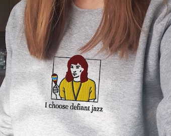 May include: A light gray crewneck sweatshirt featuring an embroidered graphic of a person with reddish-brown hair wearing a yellow shirt and holding a maraca. Below the rectangular frame, the text reads, I choose defiant jazz in black.