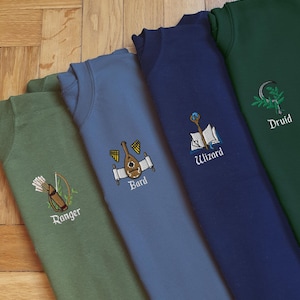 May include: Five sweatshirts in different colours with embroidered designs representing different Dungeons and Dragons character classes. The sweatshirts are arranged in a row from left to right: Ranger, Bard, Wizard, Druid. The Ranger sweatshirt is green, the Bard sweatshirt is blue, the Wizard sweatshirt is navy blue, and the Druid sweatshirt is dark green.