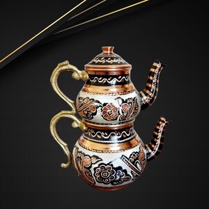 May include: A traditional Turkish double-spout teapot with intricate floral designs. The teapot is made of copper and has a black and white enamel finish. The handles are made of brass.