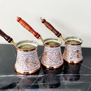 Puede incluir: Tres cafeteras de cobre con diseños florales blancos y dorados y mangos de madera. Las cafeteras están sobre una superficie de mármol negro.