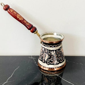Puede incluir: Una cafetera turca tradicional con cuerpo de cobre y mango de madera. La cafetera está decorada con intrincados patrones florales en negro, blanco y rosa.