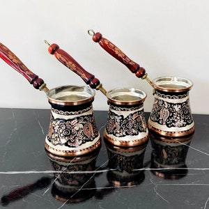 Puede incluir: Tres cafeteras turcas tradicionales con diseños florales ornamentados en negro, oro y cobre. Las cafeteras tienen mangos de madera y están dispuestas en fila sobre una superficie de mármol negro.
