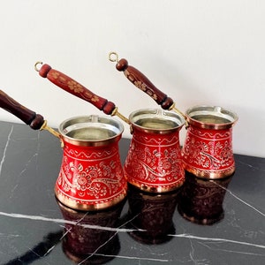 Puede incluir: Tres cafeteras turcas rojas y doradas con mangos de madera. Las cafeteras tienen un diseño floral.