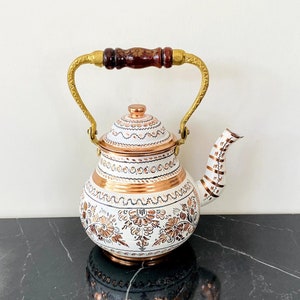 Puede incluir: Tetera de esmalte blanco con asa dorada y detalles en cobre. La tetera tiene un diseño floral y está sobre una superficie de mármol negro.