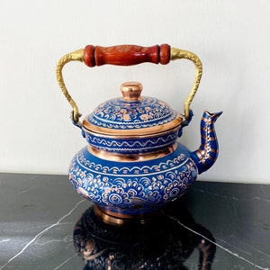 Puede incluir: Una tetera azul y cobriza con intrincados diseños florales. La tetera presenta un asa de madera con detalles dorados y una tapa a juego. El diseño incluye un pico vertedor y un cuerpo redondeado.