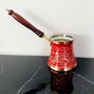 Puede incluir: Cafetera turca roja y dorada con mango de madera. La cafetera tiene un diseño floral.