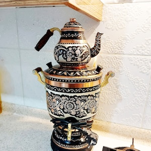 May include: A copper and black samovar with floral patterns and a matching teapot. The samovar features a spigot, handles, and a black base. The teapot has a wooden handle and a decorative spout. The kitchen appliance is on a countertop.