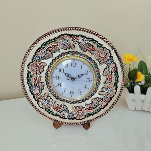 May include: A white clock with black hands and Roman numerals is set into a copper plate with a decorative floral design in shades of red, green, blue, and gold. The clock is on a wooden stand.
