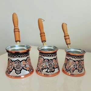 Puede incluir: Tres cafeteras turcas de cobre con mangos de madera y diseños florales intrincados. Las cafeteras están sobre una superficie blanca.