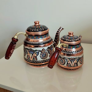 Puede incluir: Dos teteras de cobre con intrincados diseños florales pintados en negro, blanco, azul y naranja. Las teteras tienen asas de madera y están hechas en Turquía.