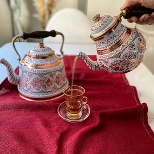 Puede incluir: Dos teteras blancas con intrincados diseños florales y detalles en cobre. La tetera de la derecha está vertiendo té en una taza de vidrio transparente con un platillo. Las tazas de té y las teteras están sobre un mantel rojo.