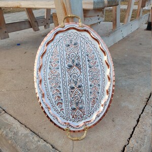 May include: An oval copper serving tray with a decorative floral pattern in black and copper. The tray has two gold handles.