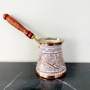 Puede incluir: Una cafetera de cobre con un mango de madera. La cafetera tiene un cuerpo blanco con diseños florales dorados y una base y borde de cobre. El mango es de color rojo oscuro con detalles dorados. Mide unos 15 cm de alto.