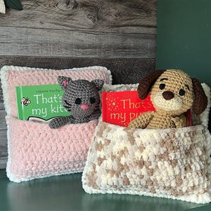Op de afbeelding: Twee gehaakte kussenbedden met knuffels erin. Het roze kussenbed heeft een grijs knuffelkatje en een boek met de titel "That's my kitten". Het beige kussenbed heeft een bruine knuffelhond en een boek met de titel "That's my puppy".