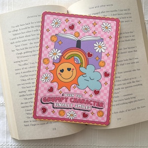 May include: A pink and yellow illustration with a rainbow, a sun with a smiley face, a cloud, and a closed book. The text "When the Grumpy Character Finally Smiles" is written in a pink and yellow font.