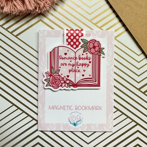 May include: A pink and red magnetic bookmark with a book design and the text "Romance books are my happy place."  The bookmark has a floral design with pink roses and white flowers. The bookmark is on a pink and white background.