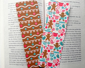 May include: Two Christmas-themed bookmarks. One bookmark features a gingerbread cookie pattern with brown, white, and pink accents. The other bookmark features a white background with a pattern of gingerbread cookies, mugs of hot chocolate, candy canes, and mittens in pink, blue, and white.