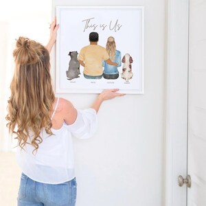 May include: A white framed print with the text "This is Us" and a watercolor illustration of a couple and two dogs. The couple is standing with their backs to the viewer, and the dogs are sitting in front of them. The names of the couple and dogs are written below the illustration.
