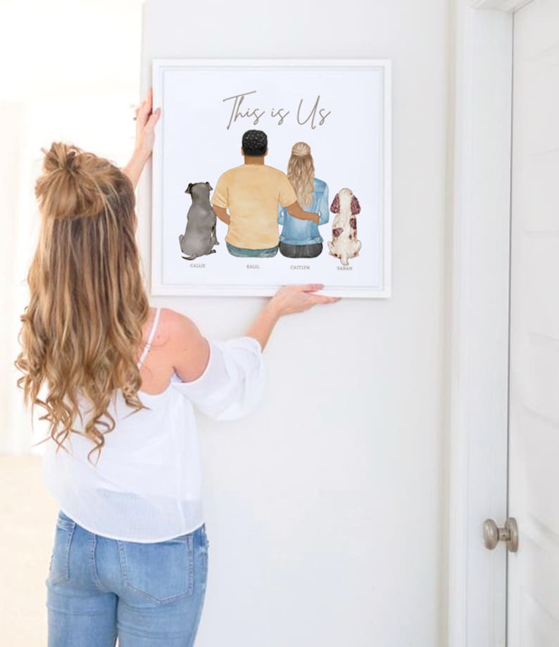 May include: A white framed print with the text "This is Us" and a watercolor illustration of a couple and two dogs. The couple is standing with their backs to the viewer, and the dogs are sitting in front of them. The names of the couple and dogs are written below the illustration.