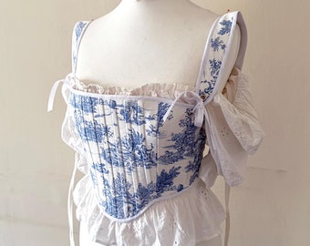 May include: A white corset with blue and white toile pattern, featuring a ruffled trim and adjustable straps. The corset is on a white dress form.