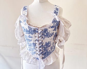 May include: A white and blue toile patterned corset with white lace trim and straps. The corset has a lace-up back and is tied with a white ribbon.