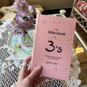 May include: A pink book titled "the little book of 3's" with the text "Prompts for play, inspiration, and self-discovery." and the author's name "Julianne Ishler".