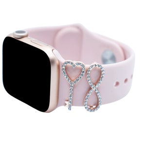 May include: A pink silicone watch band with a rose gold watch face and two charms. One charm is a heart shape and the other is an infinity symbol. Both charms are covered in clear rhinestones.