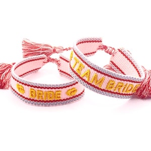 May include: Two pink and red friendship bracelets with gold embroidery. One bracelet says "Team Bride" and the other says "Bride".