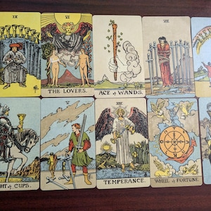 May include: A tarot card spread with ten cards, including the Knight of Cups, The Lovers, Ace of Wands, Temperance, Wheel of Fortune, and others. The cards are on a wooden surface.