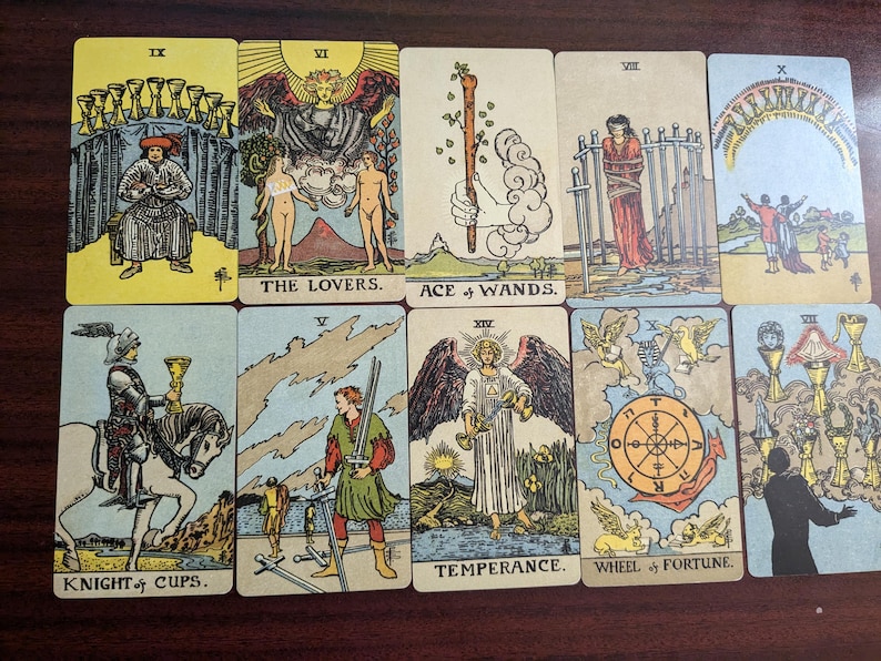 May include: A tarot card spread with ten cards, including the Knight of Cups, The Lovers, Ace of Wands, Temperance, Wheel of Fortune, and others. The cards are on a wooden surface.