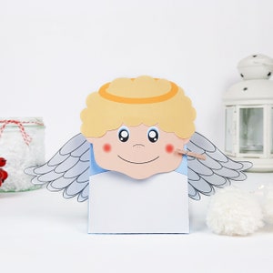May include: A white paper gift bag with a smiling angel face on the front. The angel has blonde hair, a halo, and wings. The bag is decorated with a blue border and a small clothespin.
