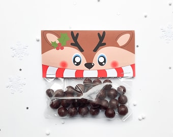 Könnte beinhalten: Ein durchsichtiger Plastikbeutel gefüllt mit dunkelbraunen runden Bonbons, versehen mit einem rechteckigen Pappreiter, der ein Rentiergesicht mit rot-weiß gestreiftem Schal und einem grünen Stechpalmenzweig mit roten Beeren zeigt.