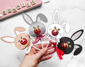 May include: Four bunny-shaped paper lollipops with colorful candy in the center. The lollipops are held in a hand and are decorated with ribbons.