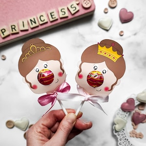 May include: Two princess-themed lollipop holders with brown hair, pink cheeks, and a crown. The lollipop holders are made of paper and have a hole in the middle for a lollipop. The lollipop holders are attached to sticks.