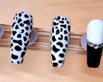 May include: Five long, oval-shaped false nails with different designs. Two nails are black, one is white with a row of rhinestones, and two are white with a black dalmatian print.