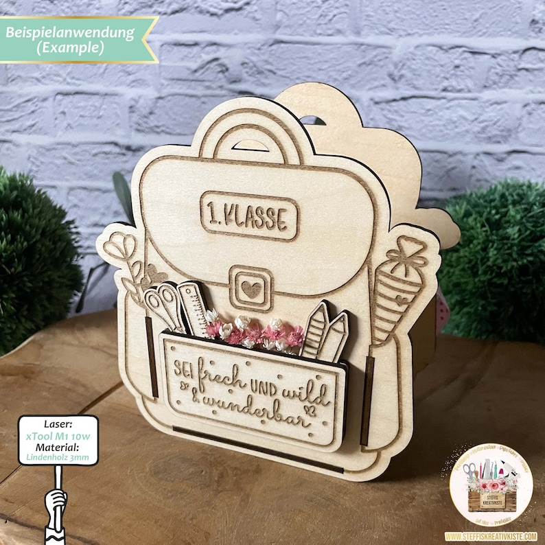 May include: A wooden cutout of a backpack with the text "1. KLASSE" and "SEI frech UND wild & wunderbar" on it. The backpack is decorated with school supplies such as pencils, scissors, and a ruler. The cutout is made from 3mm linden wood and was cut with an xTool M1 10w laser.