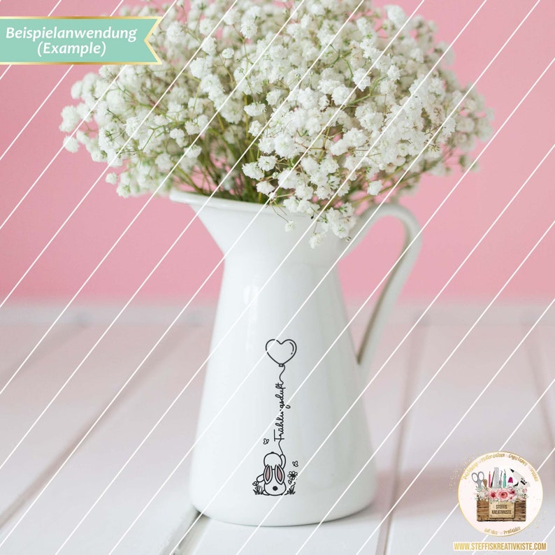 May include: A white ceramic pitcher with a black and white illustration of a bunny holding a heart-shaped balloon. The text "F&uuml;hl dich wohl" is written on the balloon. The pitcher is filled with white baby's breath flowers.