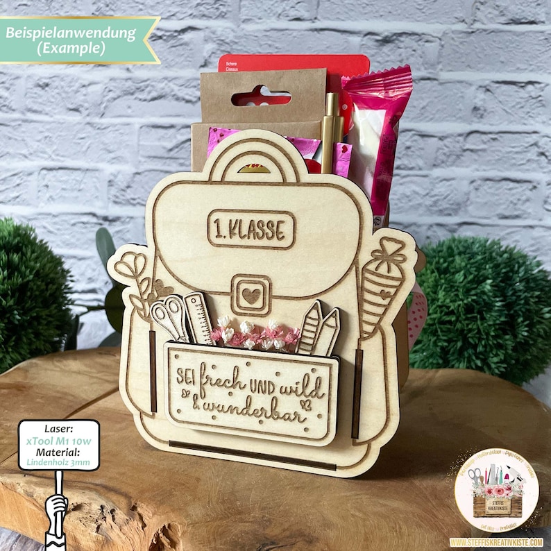 May include: A wooden backpack-shaped box with a cutout for holding school supplies. The box is decorated with the words "1. KLASSE" and "SEI frech UND wild & wunderbar".
