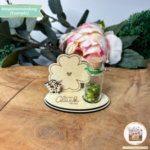 May include: A wooden clover-shaped stand with a small glass bottle filled with green glitter. The bottle has a cork top and a green ribbon. The stand has the words "Kleines Gl&uuml;ck f&uuml;r Dich" written on it.