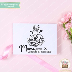 May include: A black and white illustration of a bunny rabbit holding two smaller bunnies with the text "Mama, du weist gar nicht, wie lieb wir dich haben!"  The illustration is surrounded by a wreath of flowers.