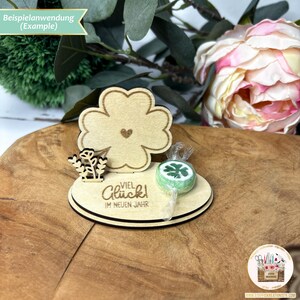 May include: A wooden four-leaf clover cutout with a heart in the center. The clover is attached to a wooden base with the text "Viel Gl&uuml;ck im neuen Jahr" (Happy New Year) and a green four-leaf clover lollipop.